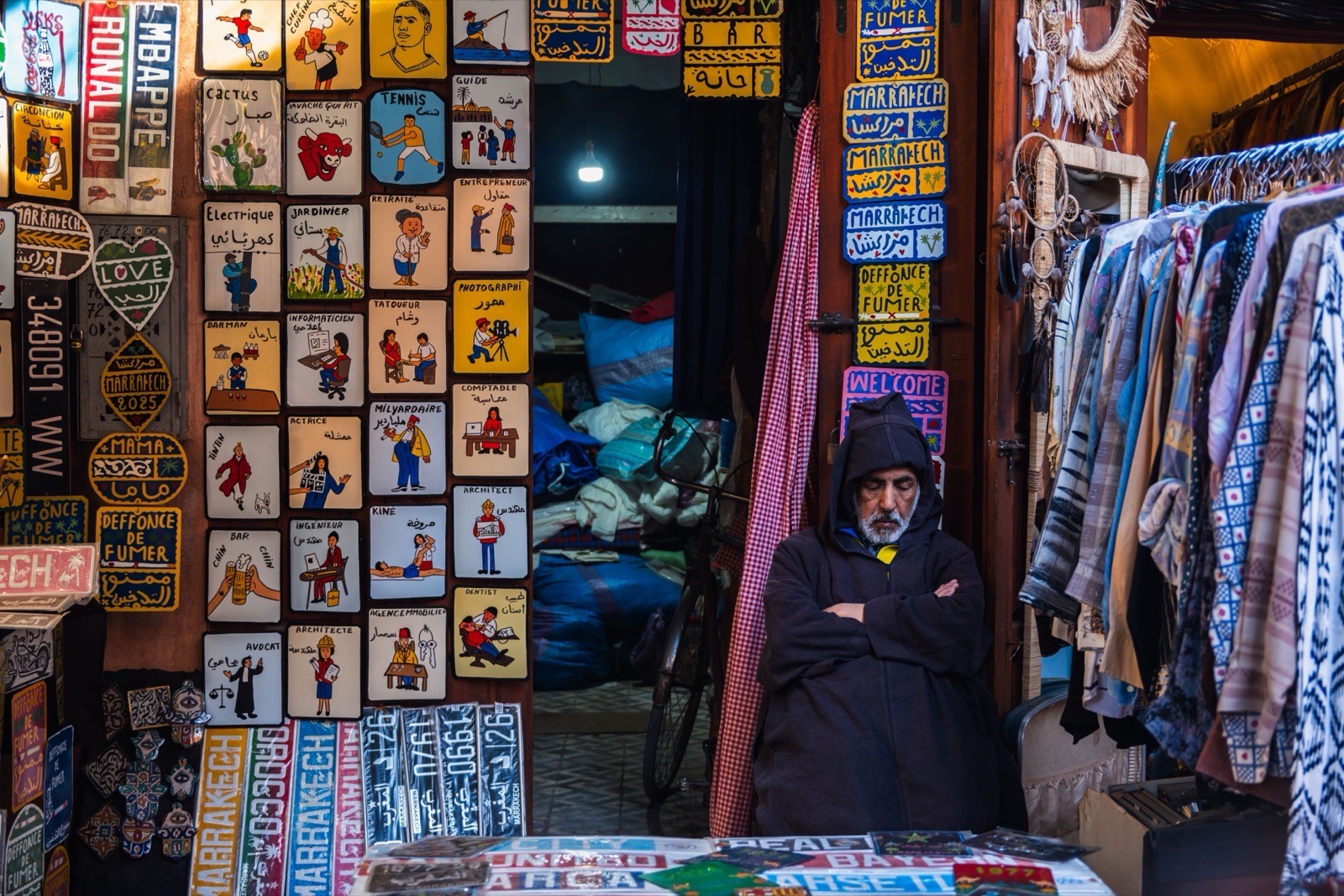 A vendor in Marrakech souk, surrounded by vibrant signs and garments, embodies local culture.
