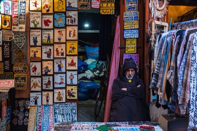 A vendor in Marrakech souk, surrounded by vibrant signs and garments, embodies local culture.